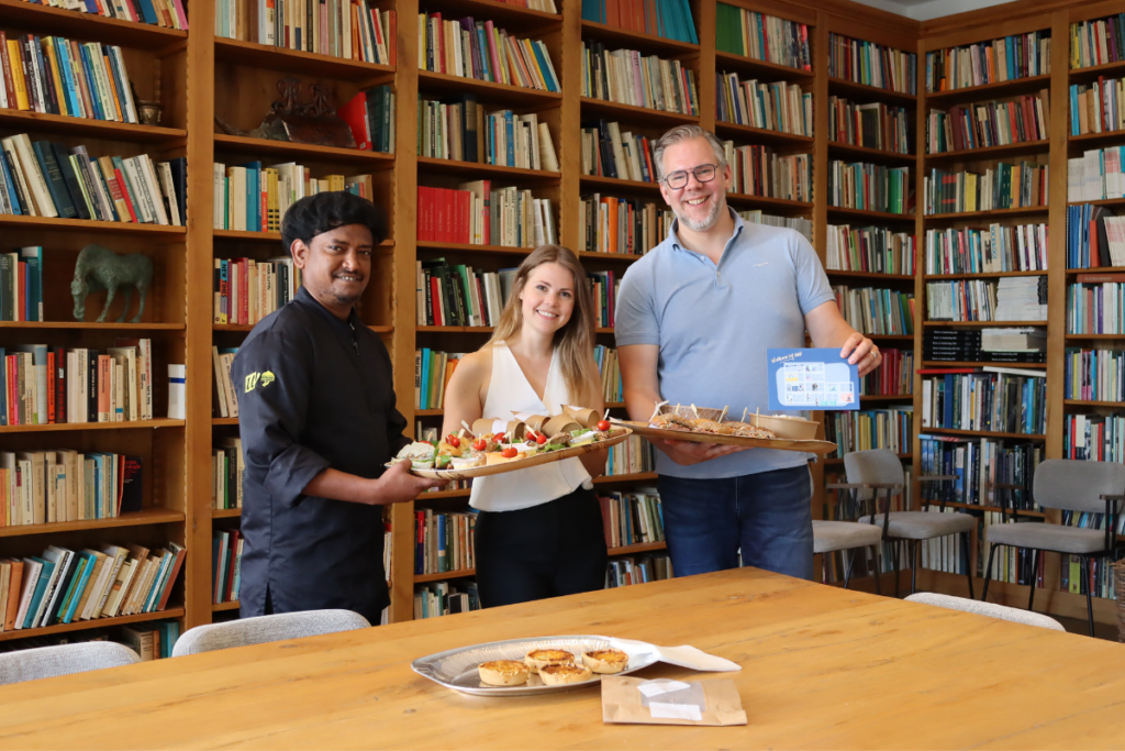 Een kok en twee presentatoren tonen schalen met luxe cateringhapjes in een sfeervolle bibliotheekruimte vol boekenkasten. Op de onderste foto staat de kok met zijn duim omhoog achter een tafel vol eten.