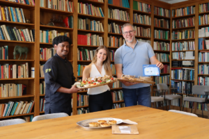 Een kok en twee presentatoren tonen schalen met luxe cateringhapjes in een sfeervolle bibliotheekruimte vol boekenkasten. Op de onderste foto staat de kok met zijn duim omhoog achter een tafel vol eten.